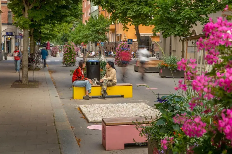 Two people talking on colorful benches surrounded by flowers on a car-free summer street.
