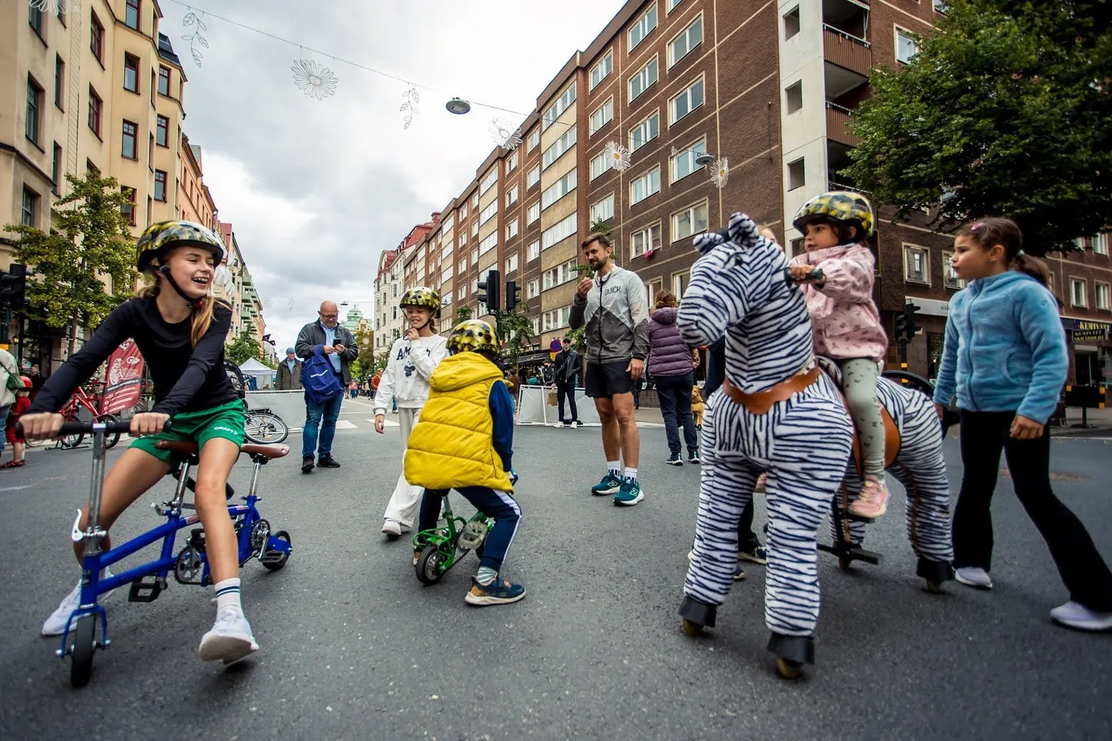 Barn som leker på en bilfri gata: en skrattande flicka på en liten cykel, en pojke i gul väst på en minicykel och ett litet barn som rider på en åkleksak formad som en zebra, medan vuxna tittar på.