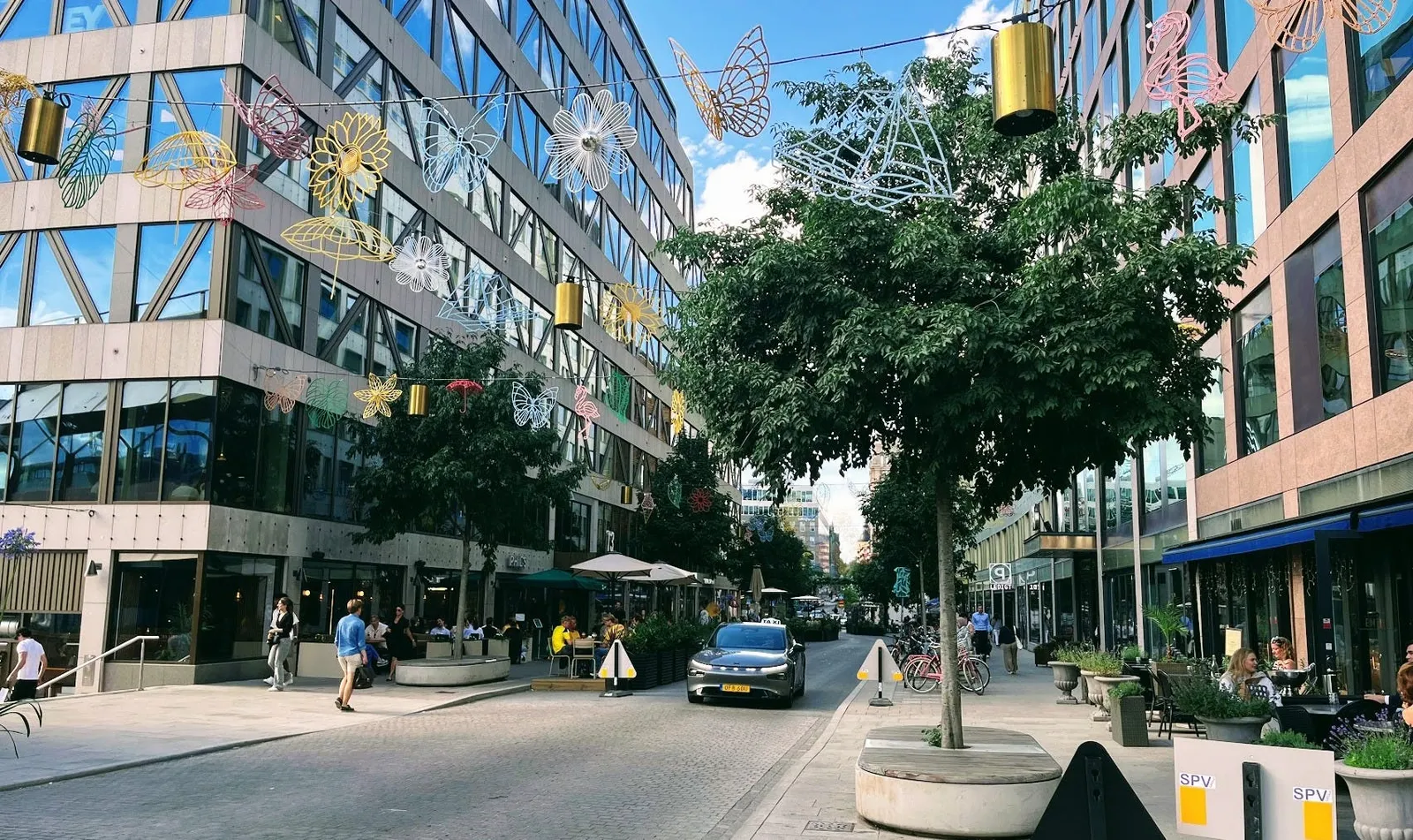 Malmskillnadsgatan på sommaren: dekorationer formade som fjärilar, blommor och flamingor hänger över gatan, och en taxi kör mitt i gatan mellan träd, uteserveringar och gående.