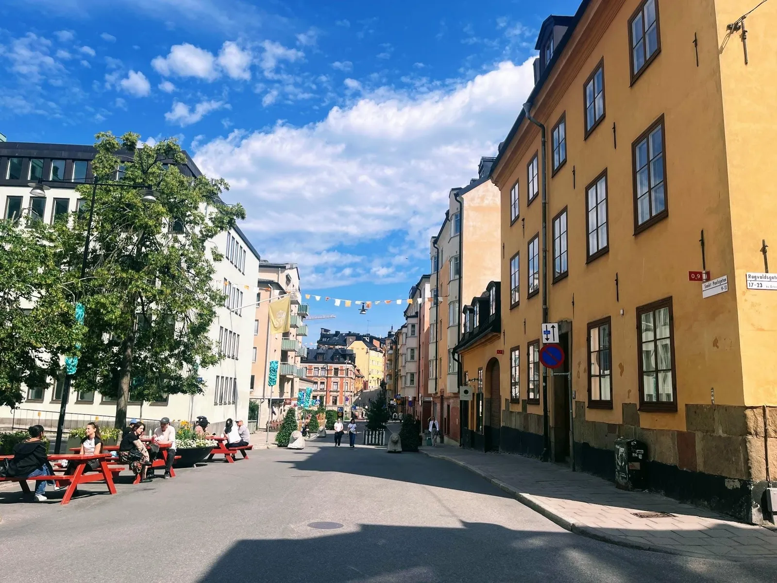 Sankt Paulsgatan på Södermalm en solig dag: röda picknickbord med folk ute i körbanan, vimplar över gatan, planteringar och gula och ockrafärgade fasader.