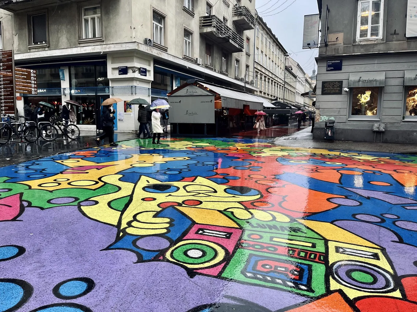 A street corner in central Zagreb where the whole roadway is covered in a colourful mural, with people walking under umbrellas.