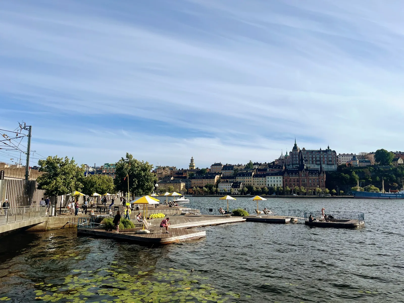 Munkbrohamnen: wooden bathing decks with yellow parasols floating on the water, with Södermalm's skyline rising behind.