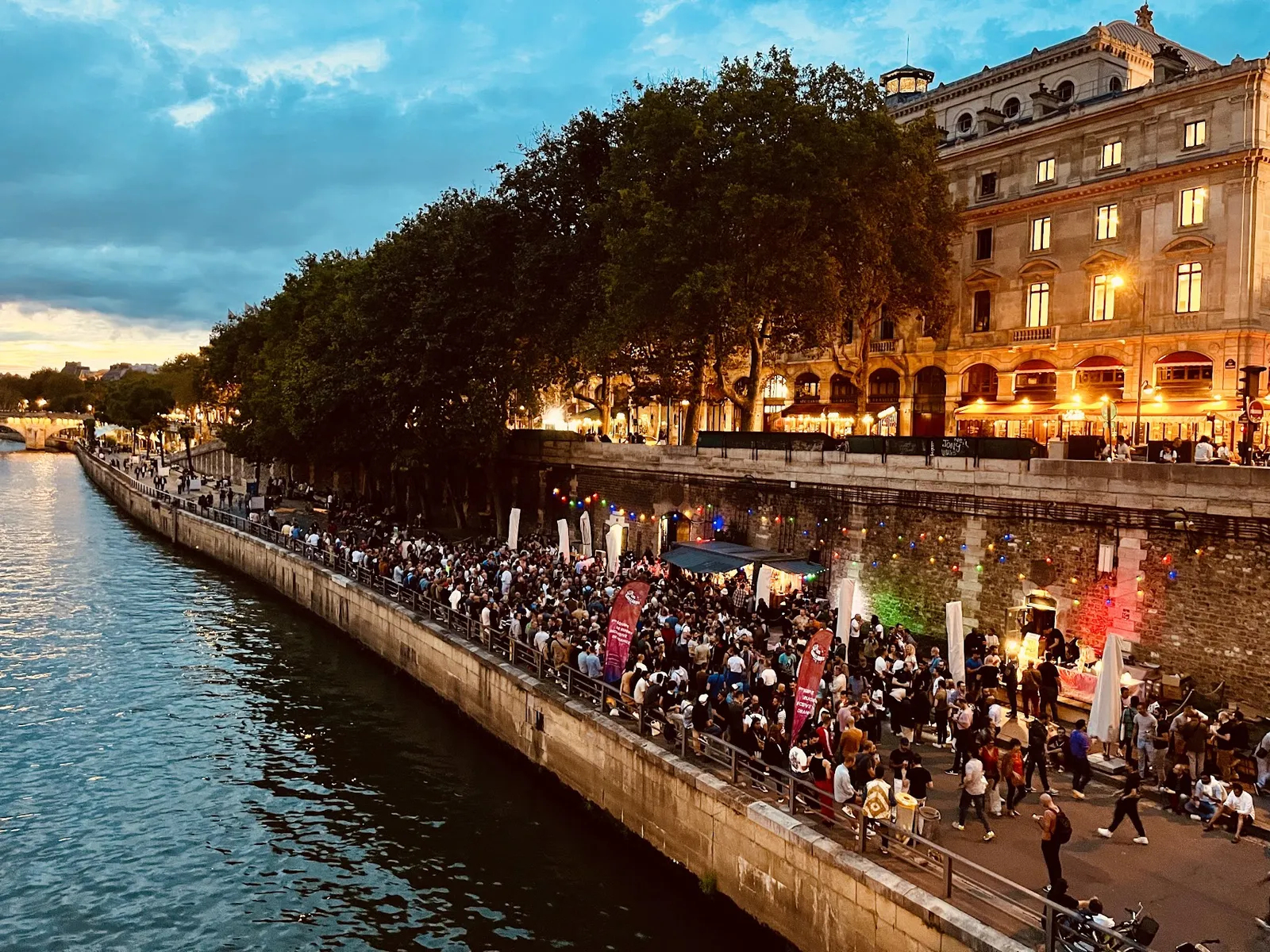Paris Plages at dusk: crowds gathered along the Seine quayside under trees and string lights, with cafés glowing in the background.